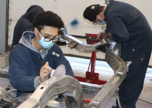 Three Automotive Trades students, wearing eyeglasses, protective face masks, and navy blue coveralls with a Capital Region BOCES logo on the front pocket, use tools to sand the body of a 1967 Corvette in a garage at the Career & Technical Education school.