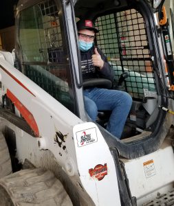 Capital Region BOCES Construction and Heavy Equipment student Remington Lawyer, wearing a black ball cap, protective eyewear, a blue protective face mask, black hoodie and jeans, sits in a piece of heavy machinery and gives a thumbs up.