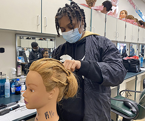 A high-school age student, who has dark brown braids and is wearing a black protective jacket and light blue face mask, styles the hair of a manequin in a Capital Region BOCES Cosmetology classroom.