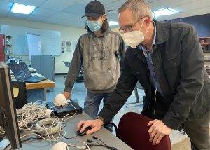 A student, wearing a black ball cap, blue protective face mask and grey sweatshirt, and a teacher, with short hair, eyeglasses and a white face mask, work together at a desktop computer in a Network Cabling classroom. White electrical cables lay on the table where they are working.