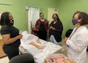 Three adult Licensed Practical Nursing students, each with dark brown hair of various lengths and floral-printed protective face masks, stand around a bed talking with a teacher, who has curly shoulder-length hair, glasses, and wears a face mask and white nurse's coat. A patient simulation mannequin lays on the bed.