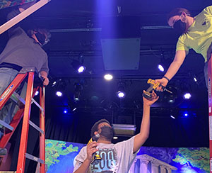 Three Entertainment Tech students, one at left standing overhead on a red ladder, one in center holding a power drill and reaching for another drill from a third student, also on a ladder at right. They are onstage at Proctors Theatre in Schenectady and surrounded by brightly colored scenery and blue light.