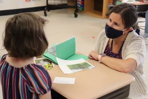 A teacher, with brown hair pulled back in a ponytail, wearing a balck face mask and white sweater, kneels to talk across a dest with an elementary-age student, with shoulder length brown hair and wearing a colorful short-sleeved striped dress. Green paper and art supplies are on the desktop.