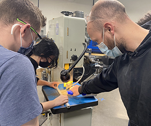 Machining program graduate and interim instructor Kody Skeals, wearing a black hoodie and protective light blue face masks, stands to the right of two students, both wearing protective eyeglasses and face masks, and instructs them on using a tabletop tool.