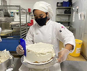 A culinary student wearing a white chefs uniform and hat and a black protective face mask spreads icing on a cake with a spatula in a Capital Region BOCES career and technical school teaching kitchen.