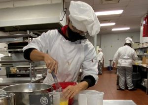 High school student Treyvon Landells, wearing a white chef's uniform and hat, eyeglasses and a protective black face mask, looks down as he uses a wisk to stir ingredientsat a workstation in a Capital Region BOCES culinary kitchen.