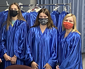 Three white Career & Technical School students, two with long brown hair and one with long blond hair, all wearing protective face masks and bright blue graduation gowns, stand side by side in front of a rack of graduation gowns. They are preparing for the upcoming Career & Technical School graduation.
