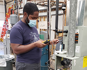 An adult learner, with a short black hair and beard who is wearing a blue t-shirt and light blue protective face mask, works with a tool and wiring in a Capital Region BOCES Heating, Ventilation, Air Conditioning and Refrigeration classroom.