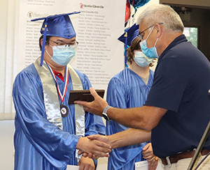 A Career & Technical school student, wearing a bright blue graduation cap and gown, eyeglasses and blue protective face mask, shakes the hand of a teacher as they receive an achievement award.