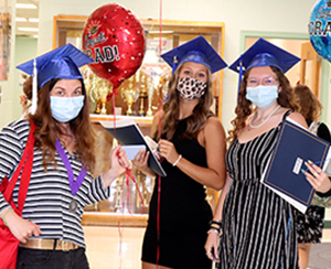 Three Schoharie Career & Technical students, all young women, wearing bright blue graduation caps and protective face coverings, stand side by side, holding red and blue balloons that read "Congratulations Grad!" They are smiling for the camera and dressed in black and white for their graduation celebration.