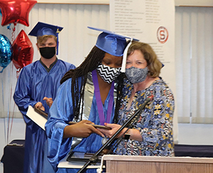 Culinary graduate Melody Francis, who has long dark brown braids and is wearing a bright blue graduation cap and gown and a white and balck zig zag patterned face covering, stands at a podium and hugs teacher Deb Toy. Toy has shoulder length light brown hair and is wearing a protective face mask and floral print top. Melody was awarded 2021 Skills USA Student of the Year. A student in blue cap and gown looks on.