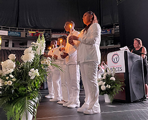 A group of adult nursing students, dressed in white, stand in a row onstage at the Albany Times Union Center. They are holding lit candles and looking out at their fellow graduates.