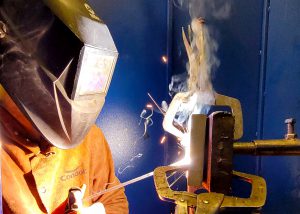An adult welding student, wearing a face shield and protective jacket, uses a tool to weld metal held in a vice.
