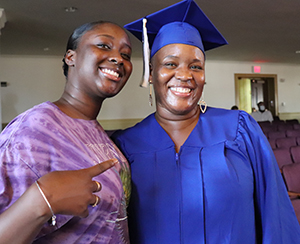 2020 High School Equivalency program graduate Katuray Korma, wearing a bright blue graduation cap and gown and rhinestone heart-shaped earrings, stands in a middle school auditorium alongside their daughter, who is wearing a purple t-dyed t-shirt with the band name Journey on its front. Both look at and smile for the camera.
