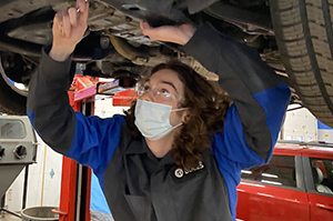 A Career & Technical School Automotive student, who has brown, shoulder-length hair and is wearing protective eyewear and bright blue and black coveralls, uses a tool to work on the underside of a vehicle.