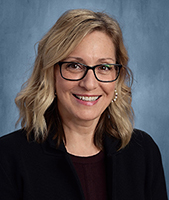 Airline Drive Academy Principal Barb Lounello, a white woman with shoulder length blond hair, glasses and a black shirt and coat, looks at and smiles for the camera.