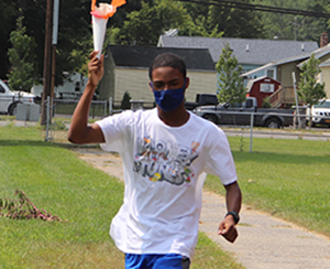 A Capital Region BOCES student, wearing a white t-shirt, bright blue shorts and carrying a paper Olympic torch, runs in a field at the Maywood School.