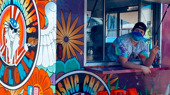 Capital Region BOCES Culinary graduate Charlie Nadler, wearing a tie-dyed t-shirt, backward black ball cap, blue face mask and making a peace sign with their fingers, looks out of the window of a brightly colored food truck with a logo that reads "Cosmic Charlie's Grateful Grill."
