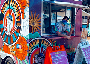 Capital Region BOCES Culinary graduate Charlie Nadler, wearing a tie-dyed t-shirt, backward black ball cap,  blue face mask and making a peace sign with their fingers, looks out of the window of a brightly colored food truck with a logo that reads "Cosmic Charlie's Grateful Grill."