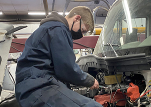 Diesel Technology student Nicholas Grock, who has short light brown hair and is wearing clear, protective eye wear, a black face mask and dark blue coveralls, works on the engine of a large truck in a repair bay at Capital Region BOCES' Career & Technical School.