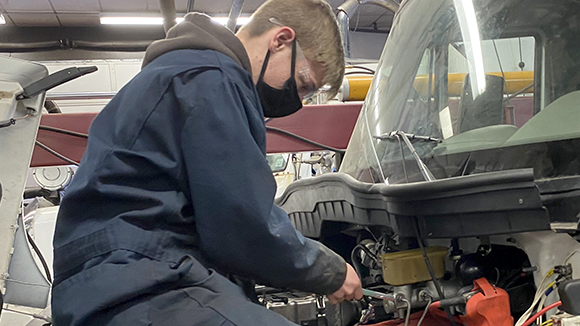 Diesel Technology student Nicholas Grock, who has short light brown hair and is wearing clear, protective eye wear, a black face mask and dark blue coveralls, works on the engine of a large truck in a repair bay at Capital Region BOCES' Career & Technical School.