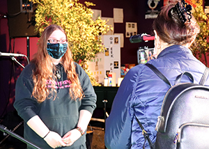 Entertainment Technology alumnus Noel Lyman, who has long light brown hair and is wearing eyeglasses, a black face mask and black hoodie with red lettering, stands on stage at Proctors Theatre in Schenectady, New York surrounded by scenery, and is interviewed on camera by a reporter from an Albany-area television station. 