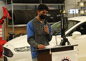 Auto Trades student Kishaun Sookdowar, who has dark brown hair and is wearing a grey and bright blue uniform top and black face covering, stands at a poduim in a front a white hybrid vehicle in an automotive garage, and speaks into a microphone.