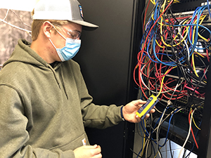  A student in the Network Cabling Technician, wearing a ball cap with a grey front and white back, clear protective eyewear, a blue protective face mask and grey hoodie, stands in front of a wall box filled with colorful wiring and takes an electrical reading with an instrument.