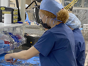 Two Sterile Processing students who stand side by side and are wearing light blue hair coverings, protective face masks and blue scrub tops, sort medical instruments at a cloth-covered metal work station.