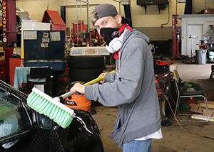 A young adult student, wearing a grey ballcap backwards, a black face covering and a grey hoodie, looks at and smiles for the camera while using a long-handled green brush to wash a black car in a wash bay at Metro Ford in Schenectady, NY.