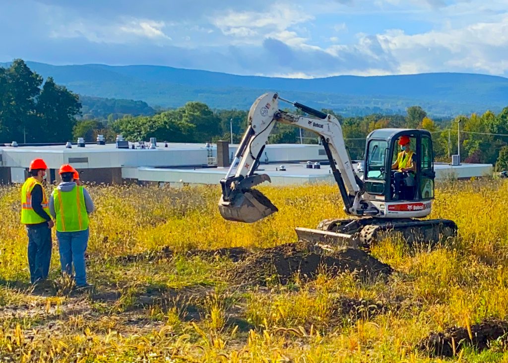 Two Heavy Equipment students, wearing bright yellow vests and orange hard hats, stand in a field of tall grass and watch a third student dig with an excavator. The roof of the Schoharie Career and Technical school and an expanse of mountains show in the distance.