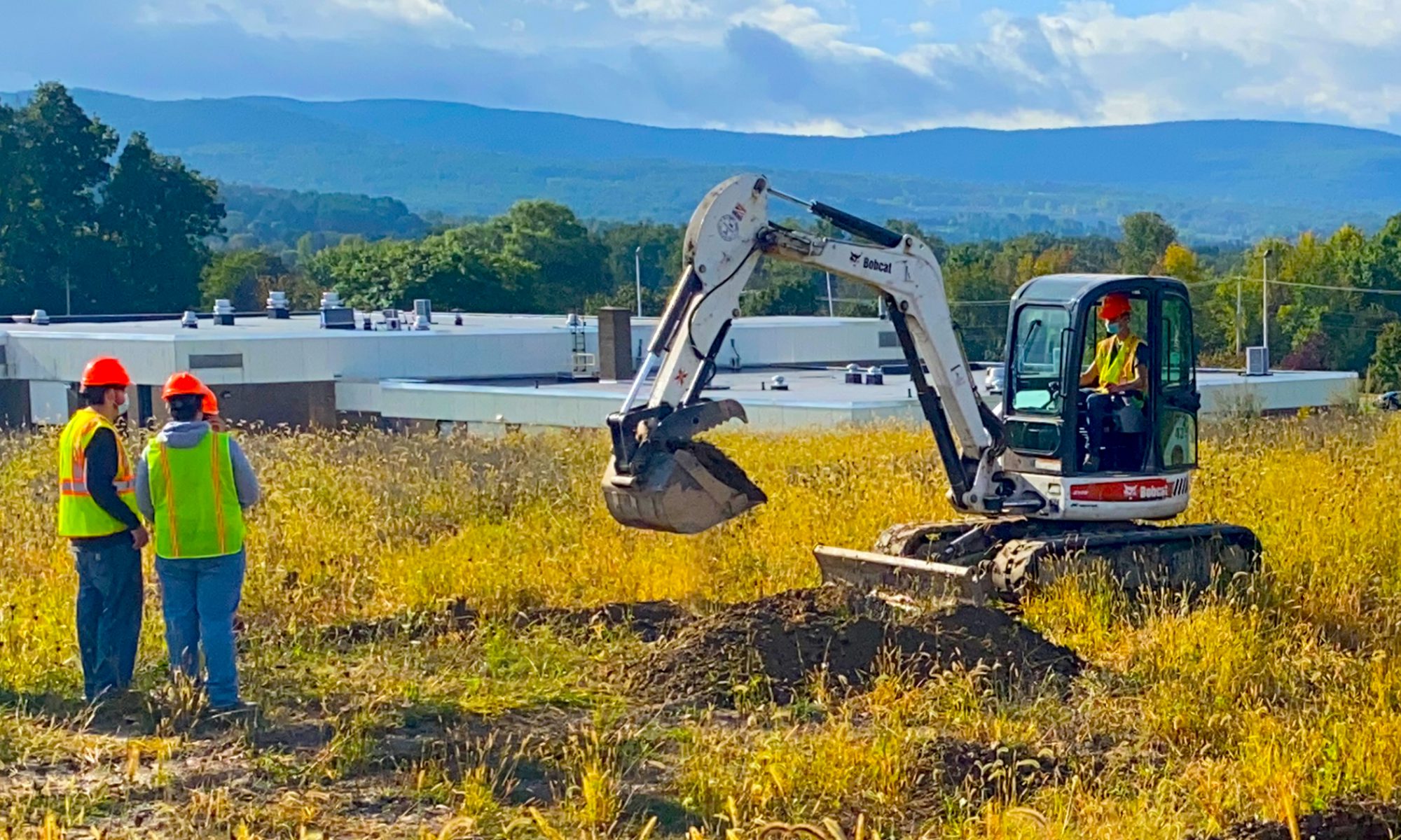 Two Heavy Equipment students, wearing bright yellow vests and orange hard hats, stand in a field of tall grass and watch a third student dig with an excavator. The roof of the Schoharie Career and Technical school and an expanse of mountains show in the distance.