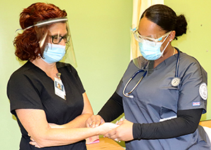 A teacher with red shoulder lenggth hair and wearing a protective face mask, clear face shield, glasses and a black scrub top has their pulse taken by an adult practical nursing student, who is wearing glasses and a protective face mask, a grey scrub top and has a stethoscope around their neck. 