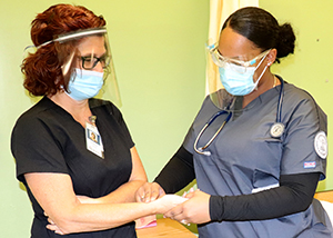 A teacher with red shoulder lenggth hair and wearing a protective face mask, clear face shield, glasses and a black scrub top has their pulse taken by an adult practical nursing student, who is wearing glasses and a protective face mask, a grey scrub top and has a stethoscope around their neck.