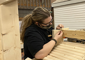 A carpentry student, who has long light brown hair held back in a ponytail, and is wearing eyeglasses, a protective face mask and black polo, joins two pieces of wood together in a Career & Technical School classroom.