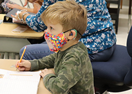 A young student, who has short blond hair, is wearing a jellybean-printed face mask with a clear window in its front, and a camo print green shirt, writes with a pencil in a lined notebook.
