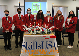 A diverse group of students, all wearing red SkillsUSA blazers, black pants, white shirts, and protective face masks stand in a row in front of a table with a large floral arrangement, lit candles and a draped in a banner that reads SkillsUSA Capital Region BOCES Career and Tech School, Albany Campus. 