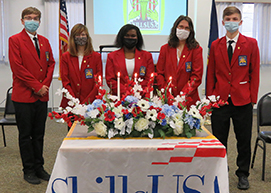  A diverse group of students, all wearing red SkillsUSA blazers, black pants, white shirts, and protective face masks stand in a row in front of a table with a large floral arrangement, lit candles and a draped in a banner that reads SkillsUSA Capital Region BOCES Career and Tech School, Albany Campus.