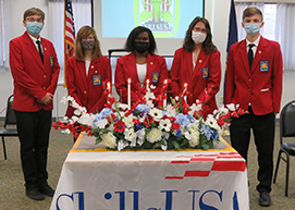 A diverse group of students, all wearing red SkillsUSA blazers, black pants, white shirts, and protective face masks stand in a row in front of a table with a large floral arrangement, lit candles and a draped in a banner that reads SkillsUSA Capital Region BOCES Career and Tech School, Albany Campus.