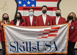  A diverse group of students, all wearing red SkillsUSA blazers, black pants, white shirts, and protective face masks stand in a row behind a banner that reads SkillsUSA. 