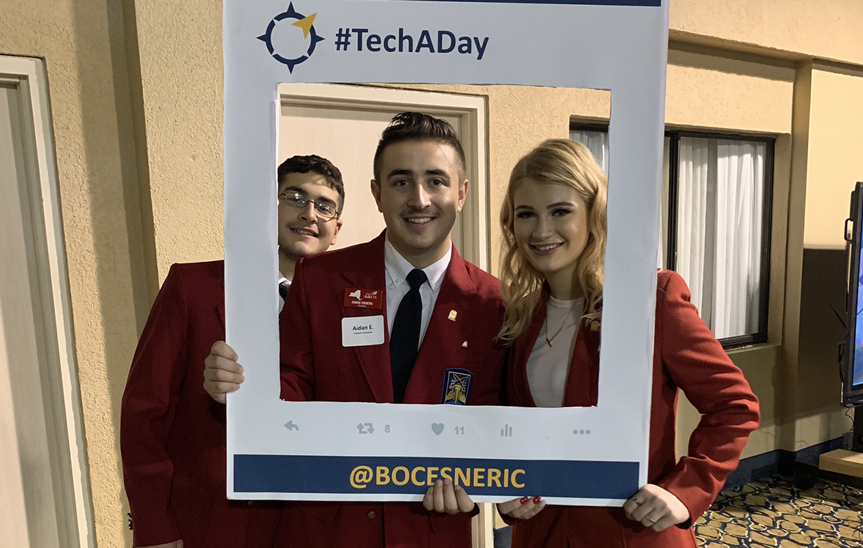 Three students wearing red SkillsUSA blazers, stand side by side and hold a large white selfie frame that reads I'm At Tech A Day. All look at and smile for the camera.