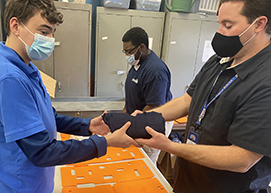 A student in the Retail and Office Services program, who has short wavy brown hair and is wearing a blue face mask and bright blue polo shirt, works with a teacher who wears a black face mask and polo, to fold an article of clothing.