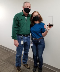 Melissa Peck, who has long, wavy red hair and is wearing eyeglasses, a black face mask and a bright blue shirt, holds a clear commitment award and stands alongside BOCES Director of Facilities Tom Mott, who wears a black protective mask and long-sleeved green shirt.