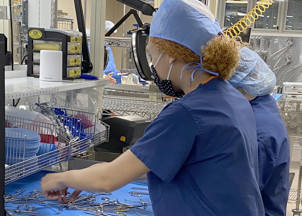 Two Sterile Processing students who stand side by side and are wearing light blue hair coverings, protective face masks and blue scrub tops, sort medical instruments at a cloth-covered metal work station.