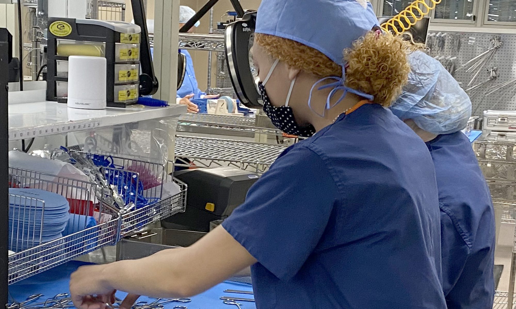 Two Sterile Processing students who stand side by side and are wearing light blue hair coverings, protective face masks and blue scrub tops, sort medical instruments at a cloth-covered metal work station.