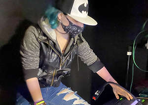 An Entertainment Tech student, who has teal blue shoulder-length hair and is wearing a black and white ball cap with a Batman logo and a black bandana print face mask, works at the sound board at Proctors Theatre in Schenectady.