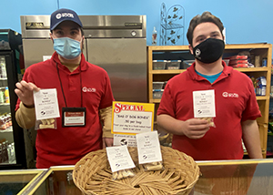 Two retail/office services students, both wearing red polo shirts with a small white Capital Region BOCES logo and face masks, stand side by side in a classroom kitchen and hold dog bones they have made to support the Mohawk Hudson Humane Society.