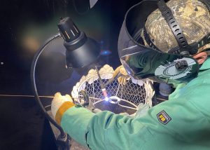A student, wearing a protective helmet and face shield and green welder's jacket, welds metal with a MIG torch in a Career and Technical School classroom.