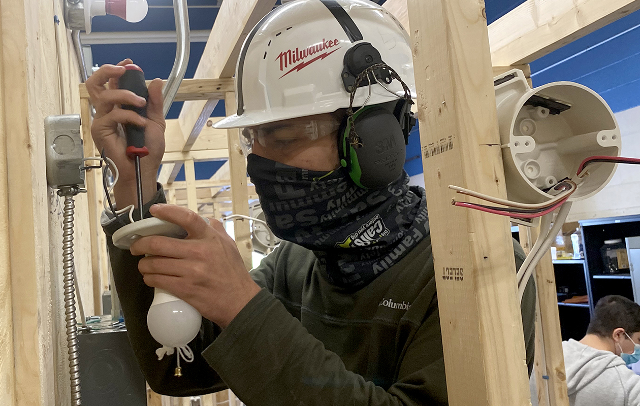 An Electrical Trades student, wearing a white hard hat, protecting ear and eyewear, a black bandana as a face mask and grey shirt, uses a screwdriver to install a light fixture in a Career and Technical School classroom.