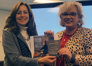 Virginia Bond, who has short wavy blond hair and is wearing eyeglasses, a red shirt and leopard print sweater, holds commitment award and photograph and stands alongside NERIC Manager for Instructional Technology Services, who has long brown hair and is wearing a white turtleneck sweater and grey jacket. Both are smiling for the camera.